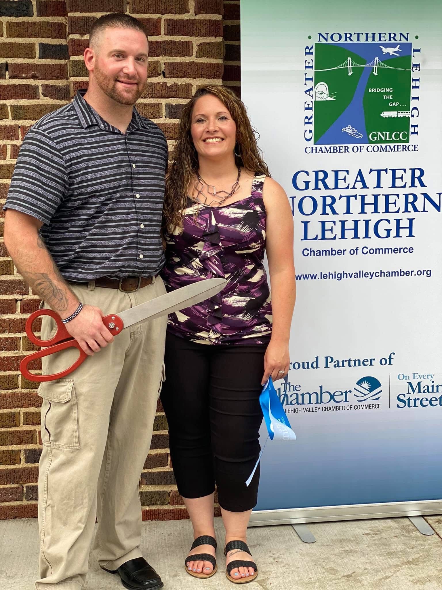 Aaron with his wife Tanya in front of a Greater Northern Lehigh Chamber of Commerce banner, holding oversized ceremonial scissors